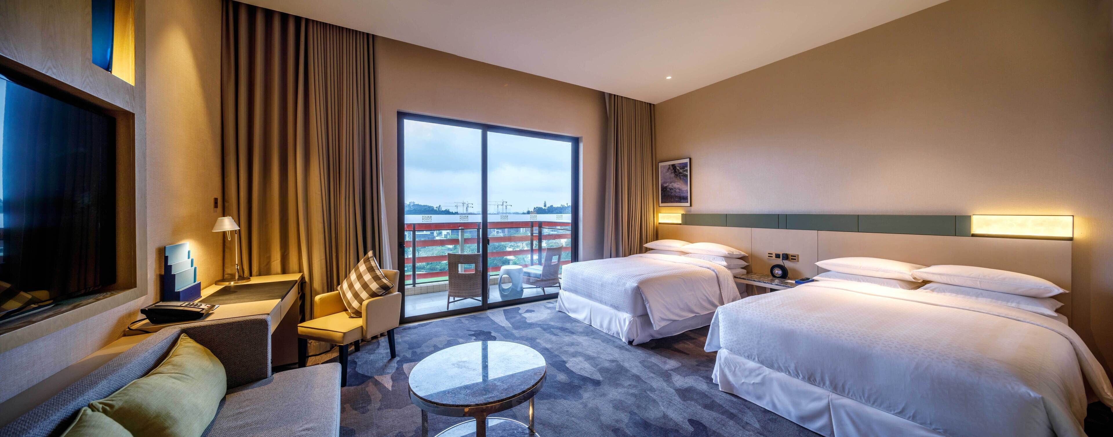 A hotel room with two beds, a sofa, a small table, a TV, and a balcony with a view of the city.