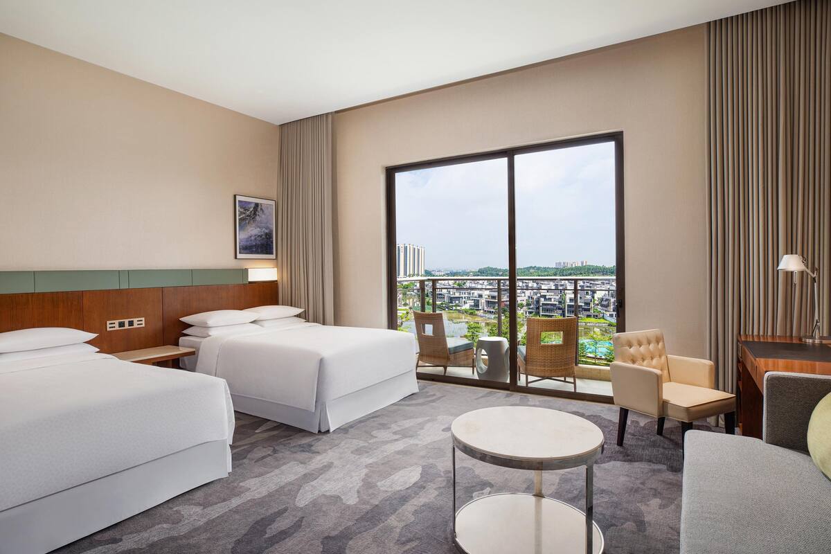 Quarto de hotel com duas camas, varanda com vista para a cidade, uma mesinha e uma cadeira.