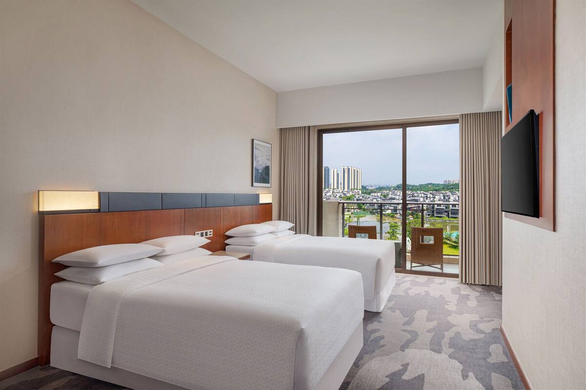 Quarto de hotel com duas camas, uma janela grande com vista para a cidade, televisão de tela plana na parede e uma varanda pequena com uma cadeira.