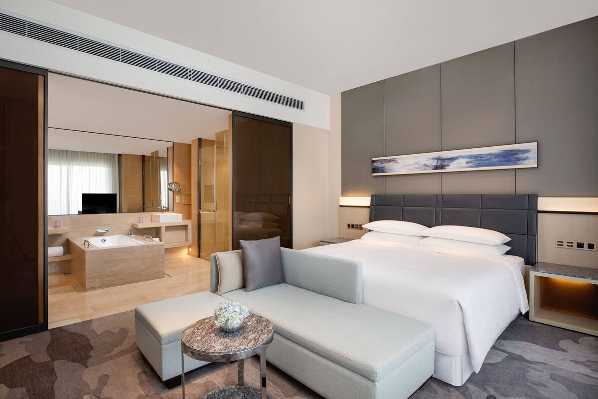 Quarto de hotel moderno com uma cama grande, um sofá, uma mesa pequena e banheiro com banheira e chuveiro.
