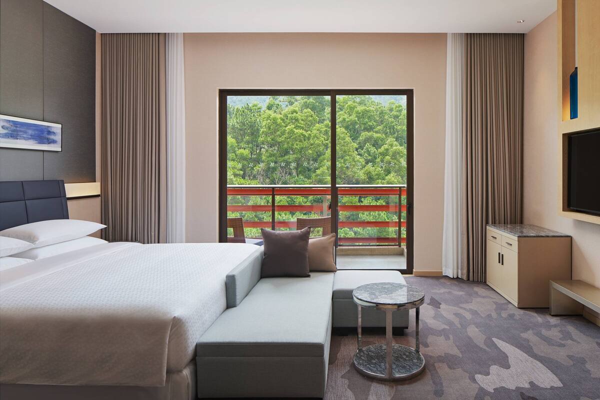 Quarto de hotel moderno com cama, sofá, televisão e varanda com vista para árvores.