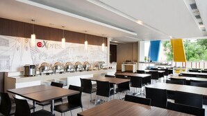 A modern dining area with multiple tables and chairs, a buffet counter with food items, and a large window with a view of greenery. - Hotel Amaris Sriwedari Solo (Surakarta)
