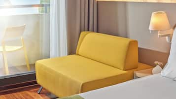 Ein Hotelzimmer mit einem gelben Sofa, einem weißen Bett, einem Nachttisch mit Lampe und Blick ins Freie durch ein Fenster.