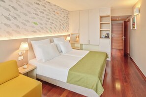 Une chambre d’hôtel avec un lit, une table de chevet avec une lampe, une petite table avec un vase, un fauteuil jaune et une armoire en bois.