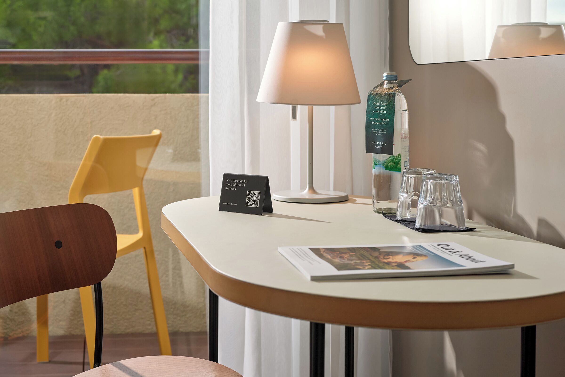 A hotel room with a table, a chair, a lamp, a minibar, and a window with a view.