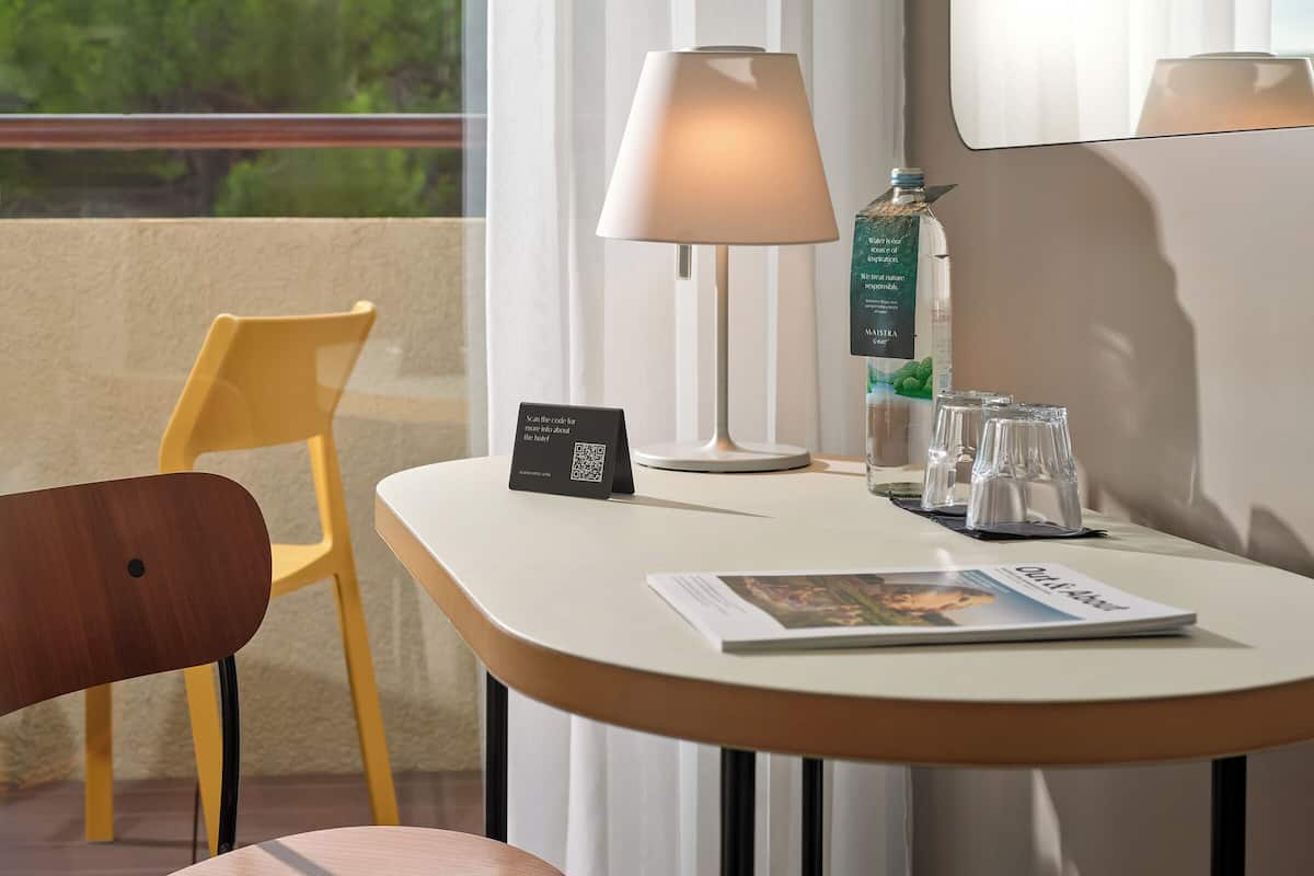 Habitación de hotel con una silla de madera, una mesa blanca con una revista, una lámpara y un minibar.
