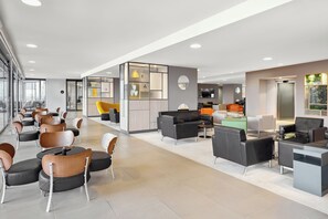 A modern hotel lobby with seating areas, a reception desk, and a kitchen in the background.