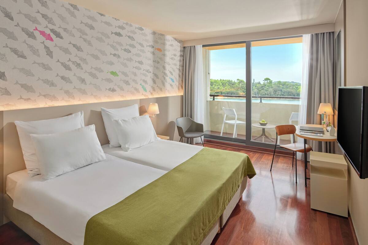 Habitación de hotel con dos camas, un balcón con vistas, un escritorio con silla y una televisión.