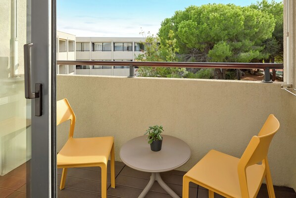 Un balcon avec deux chaises jaunes et une petite table sur laquelle est posée une plante en pot, donnant sur un immeuble et des arbres.