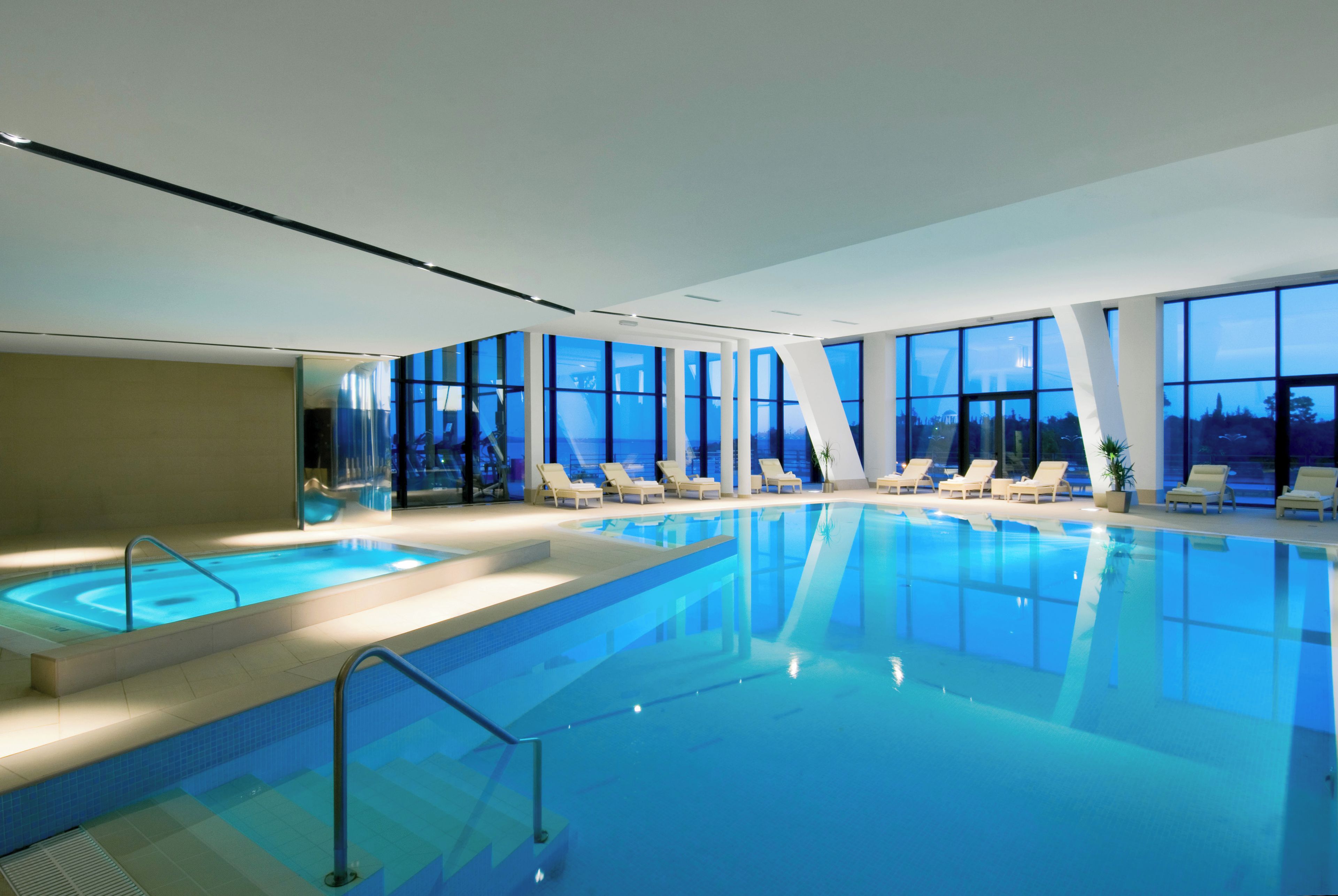 Une grande piscine intérieure à l’eau d’un bleu limpide, entourée de chaises longues et de grandes fenêtres.