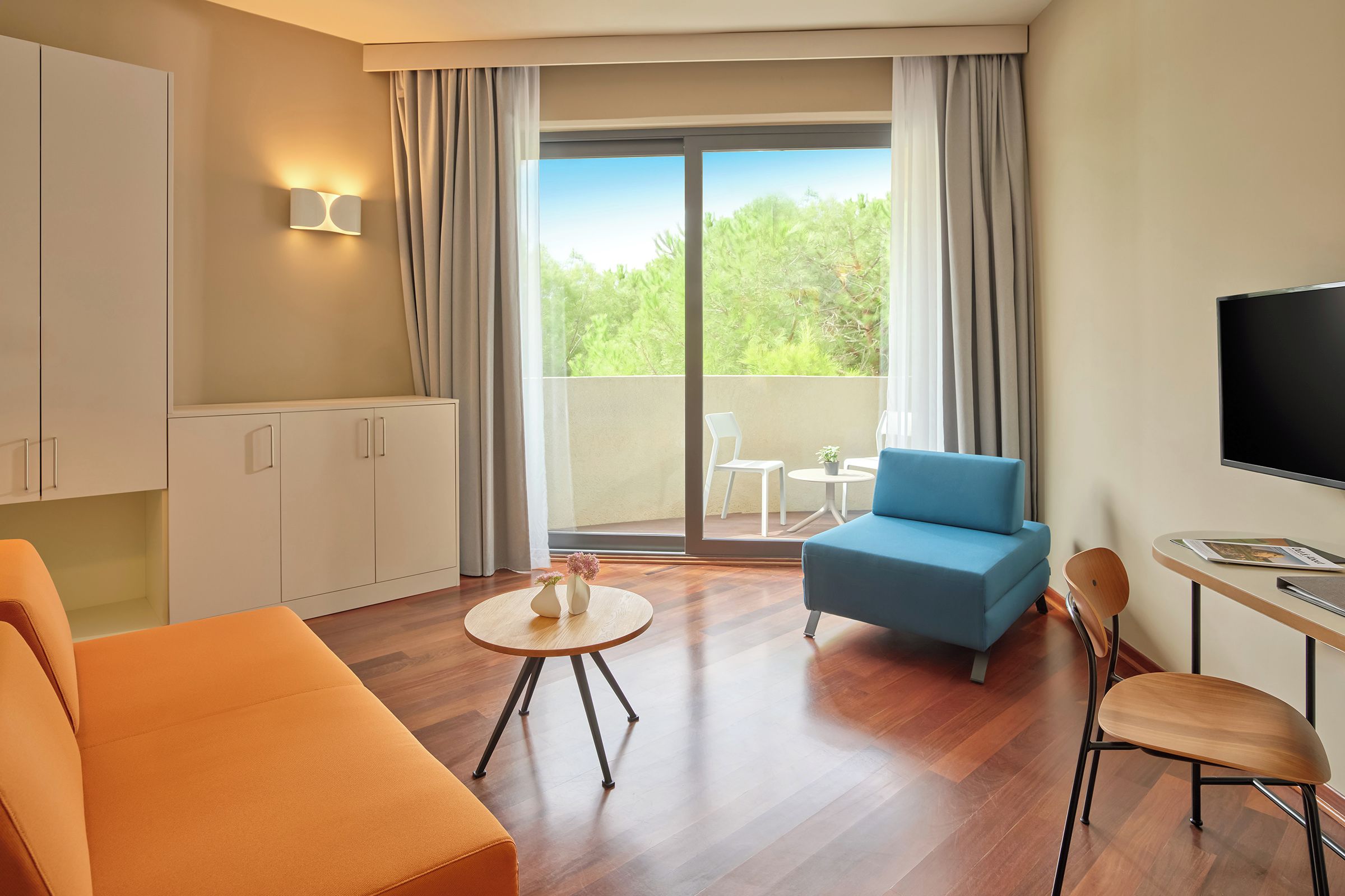 Een moderne hotelkamer met een bank, een ronde tafel, een televisie en een balkon met uitzicht op bomen.