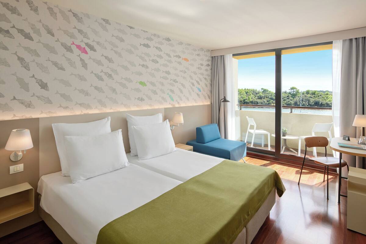 Habitación de hotel con dos camas, balcón con vistas y una pequeña zona de comedor.