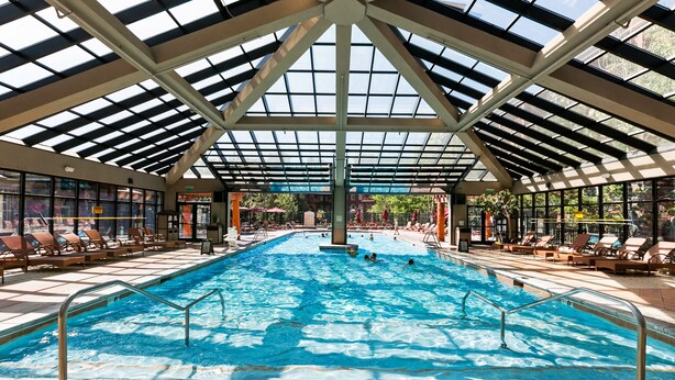 Une piscine intérieure dotée d’un toit en verre, entourée de chaises longues et recouverte d’un sol carrelé.