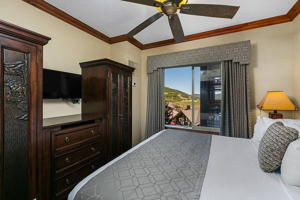 Een hotelkamer met een groot bed, een plafondventilator, een televisie, een dressoir en een raam met uitzicht op een heuvel.
