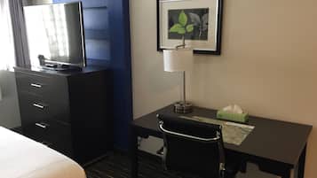 A hotel room with a bed, a desk with a chair, a television on a dresser, a framed picture on the wall, and a bedside table with a tissue box.