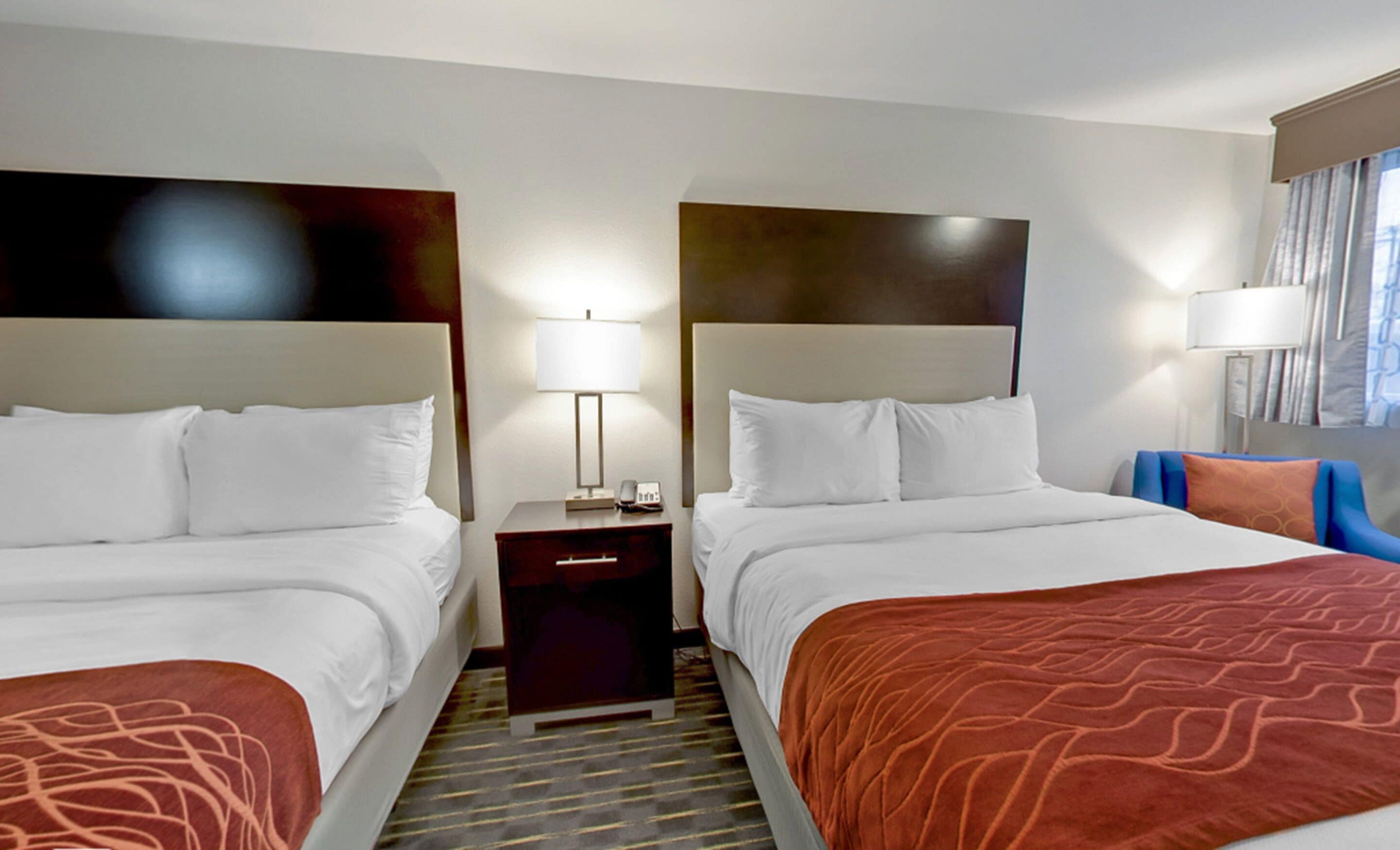 A hotel room with two beds, a nightstand, a lamp, and a chair.