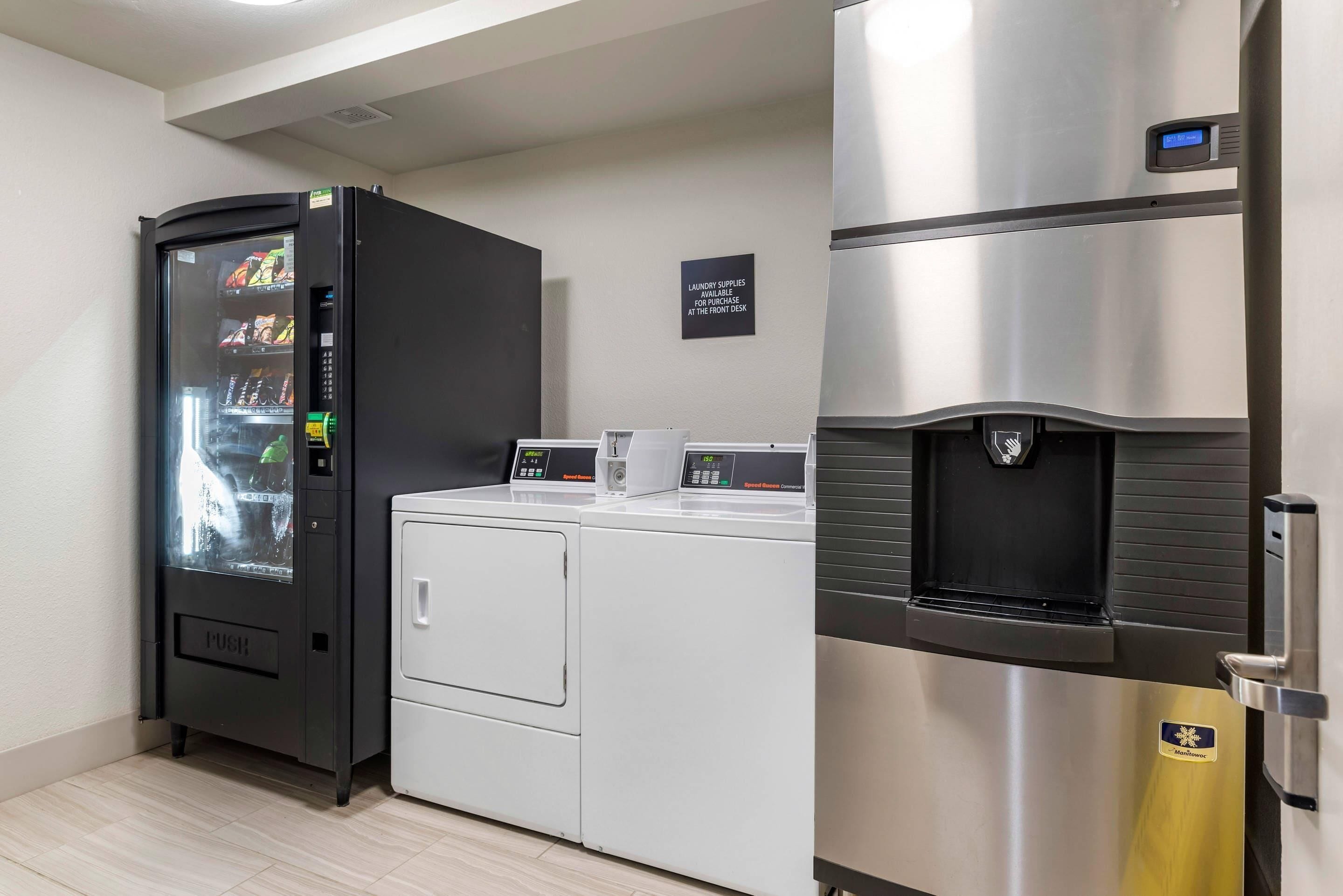 A compact kitchenette with a black vending machine, a white washing machine, and a stainless steel refrigerator.