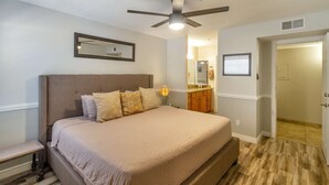 Une chambre à coucher avec un lit, un ventilateur de plafond, une table de chevet, un miroir et une salle de bain visible à travers une porte ouverte.