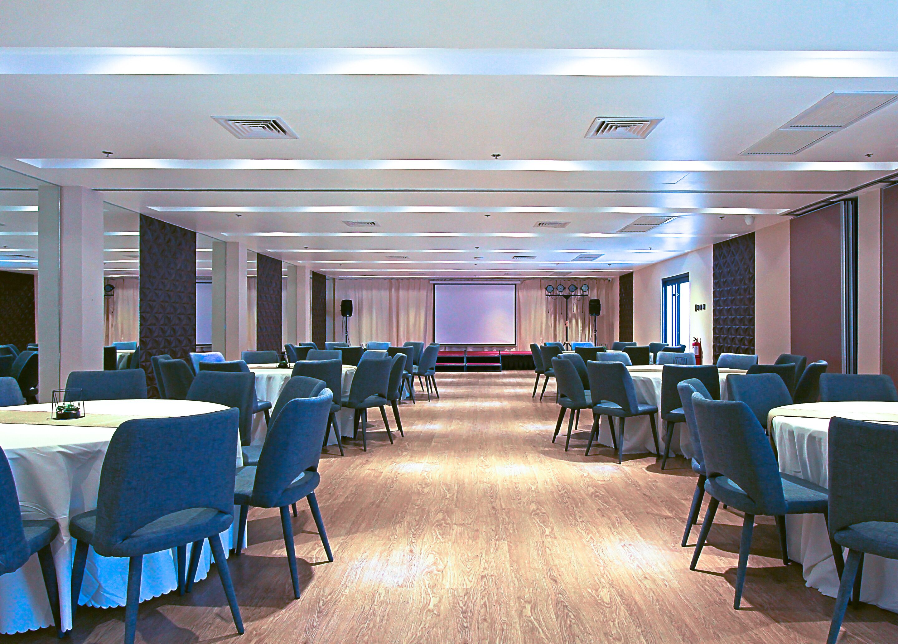 Un salón de conferencias con varias mesas y sillas, una pantalla de proyector y un escenario.