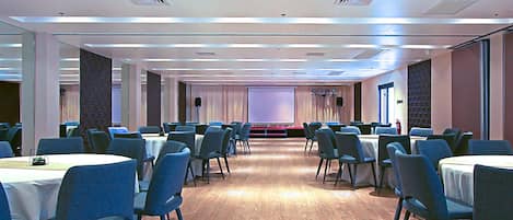 Un salón de conferencias con varias mesas y sillas, una pantalla de proyector y un escenario.
