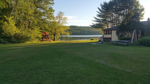 A grassy area with a small building, a swing set, and a lake in the background.