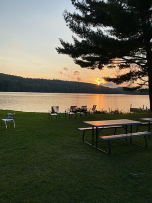 Zona de picnic junto al lago con bancos y sillas, con vistas a un lago y al atardecer de fondo.