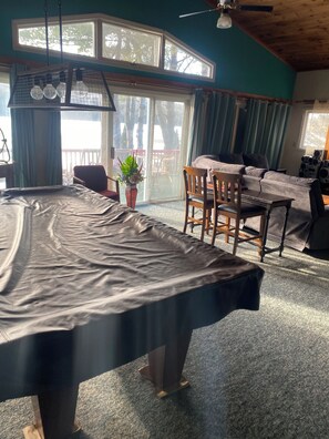Una habitación con mesa de billar, sofá y mesa de comedor. Cuenta con un amplio ventanal con cortinas, un ventilador de techo y una planta en el alféizar.