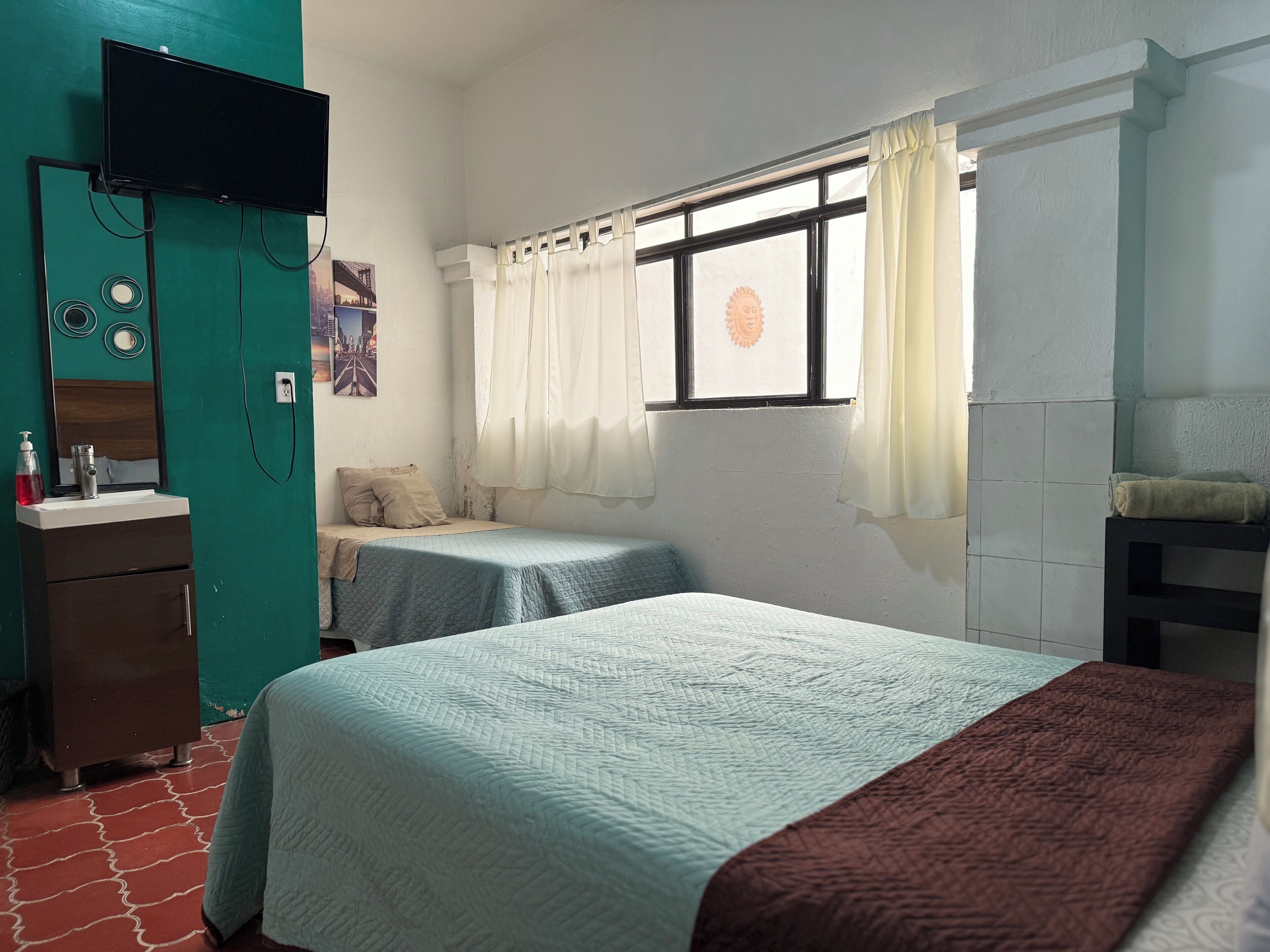 Una habitación con dos camas, un televisor montado en la pared, una ventana con cortinas, un lavamanos y un espejo.