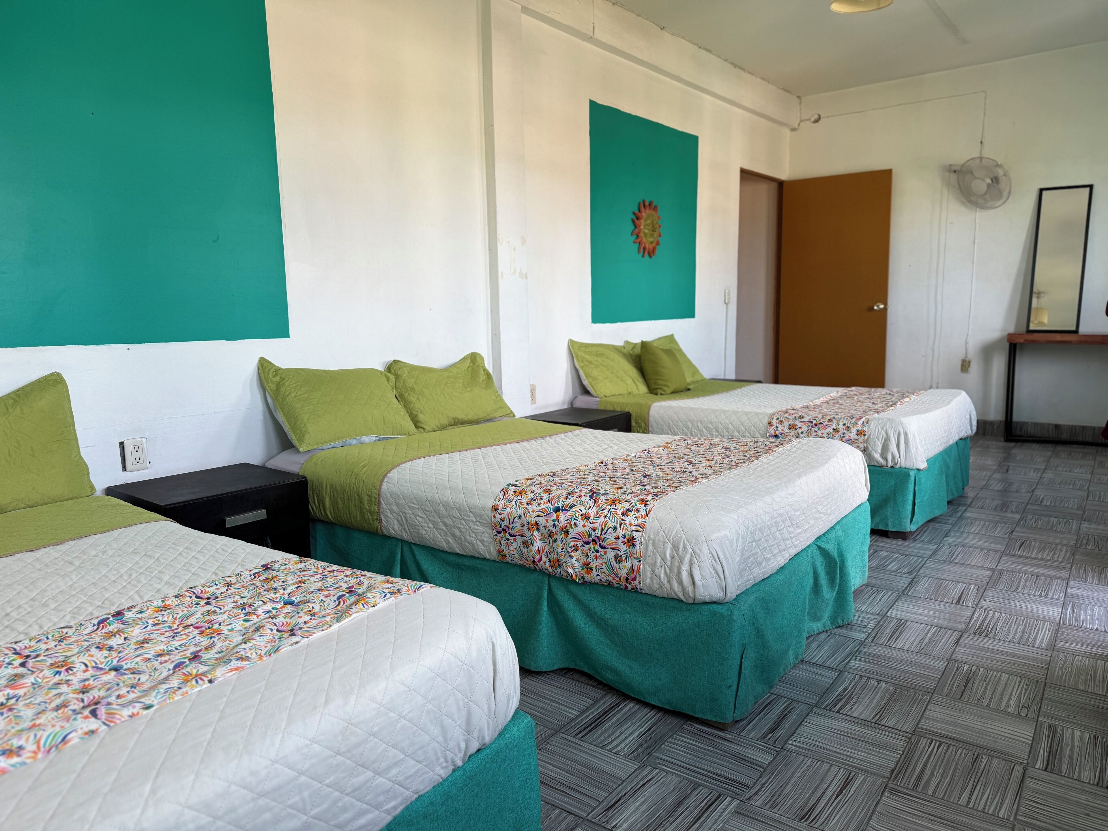 Ein Hotelzimmer mit drei Betten, jedes mit einer grünen Tagesdecke und einer farbenfrohen Blumensteck. Es gibt einen kleinen Tisch mit einem Stuhl, einen Wandventilator und eine Tür zu einem anderen Raum.