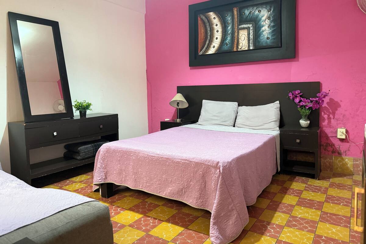 A bedroom with a bed, a nightstand, a dresser, a mirror, a wall art piece, and a potted plant.