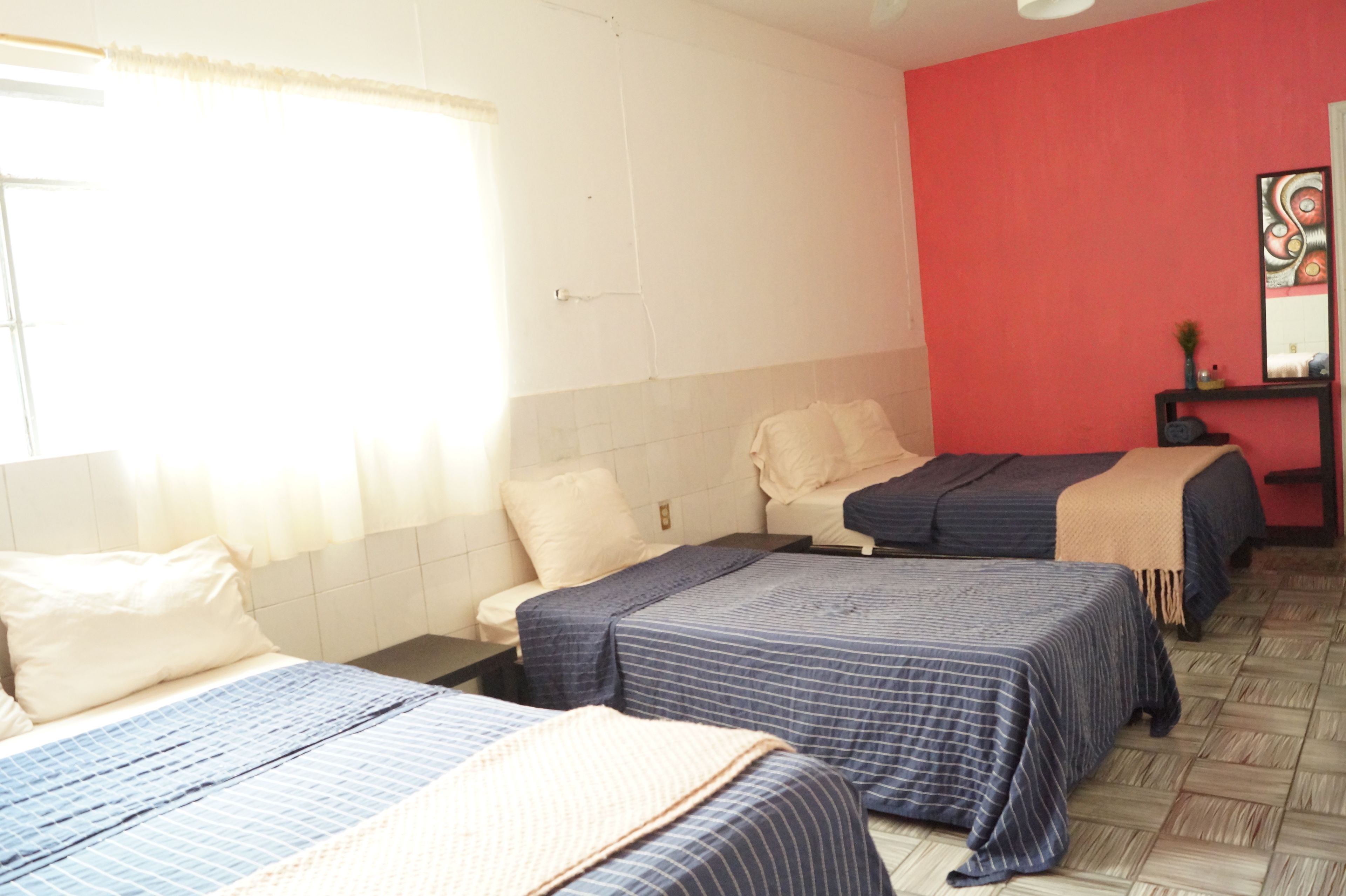 Une chambre d’hôtel avec trois lits, un mur d’accent rouge, un sol carrelé et une petite table agrémentée d’un miroir et d’objets décoratifs.
