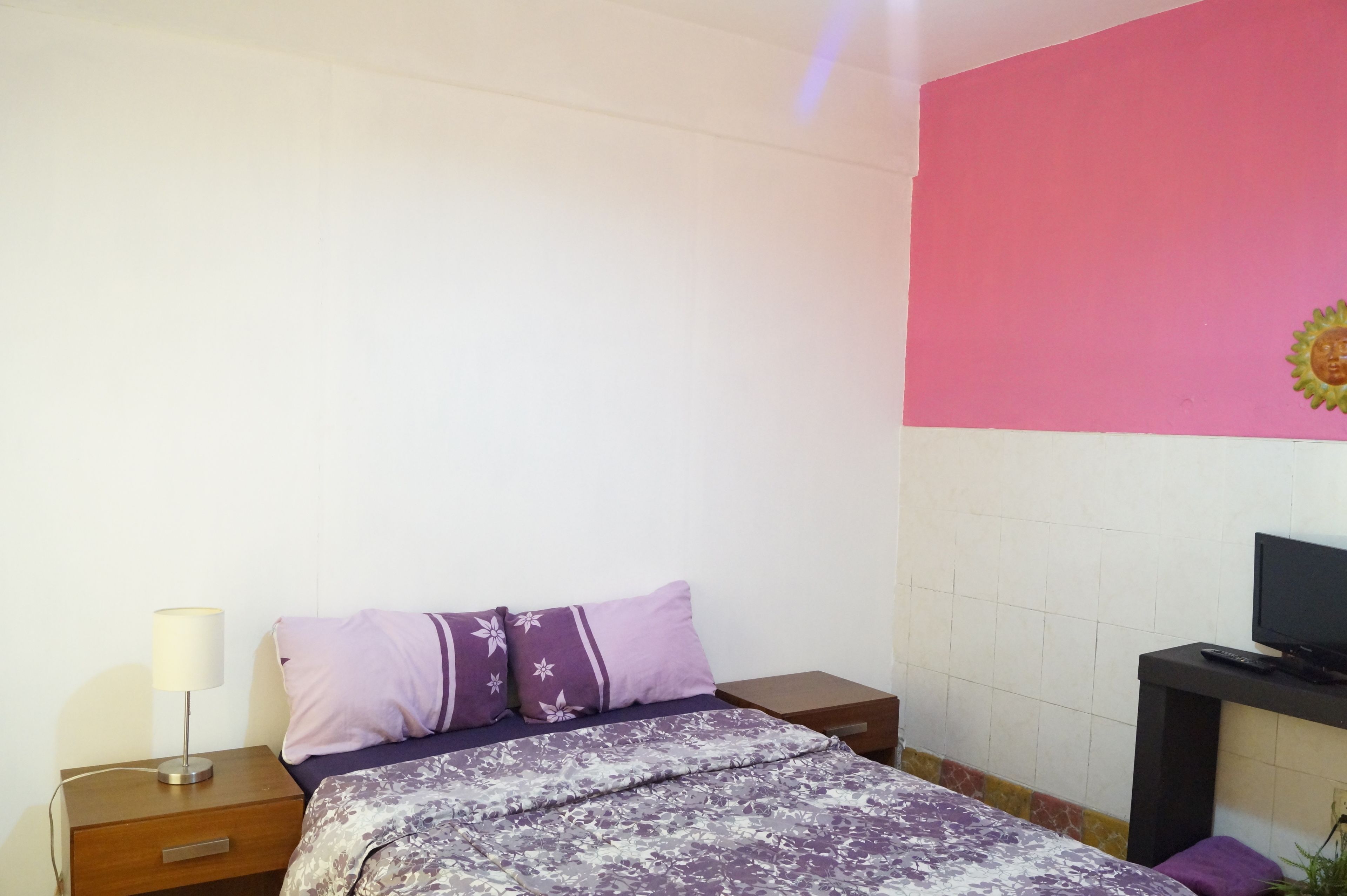 Un dormitorio con cama, mesita de noche, lámpara, televisión y un muro decorado con un motivo solar.