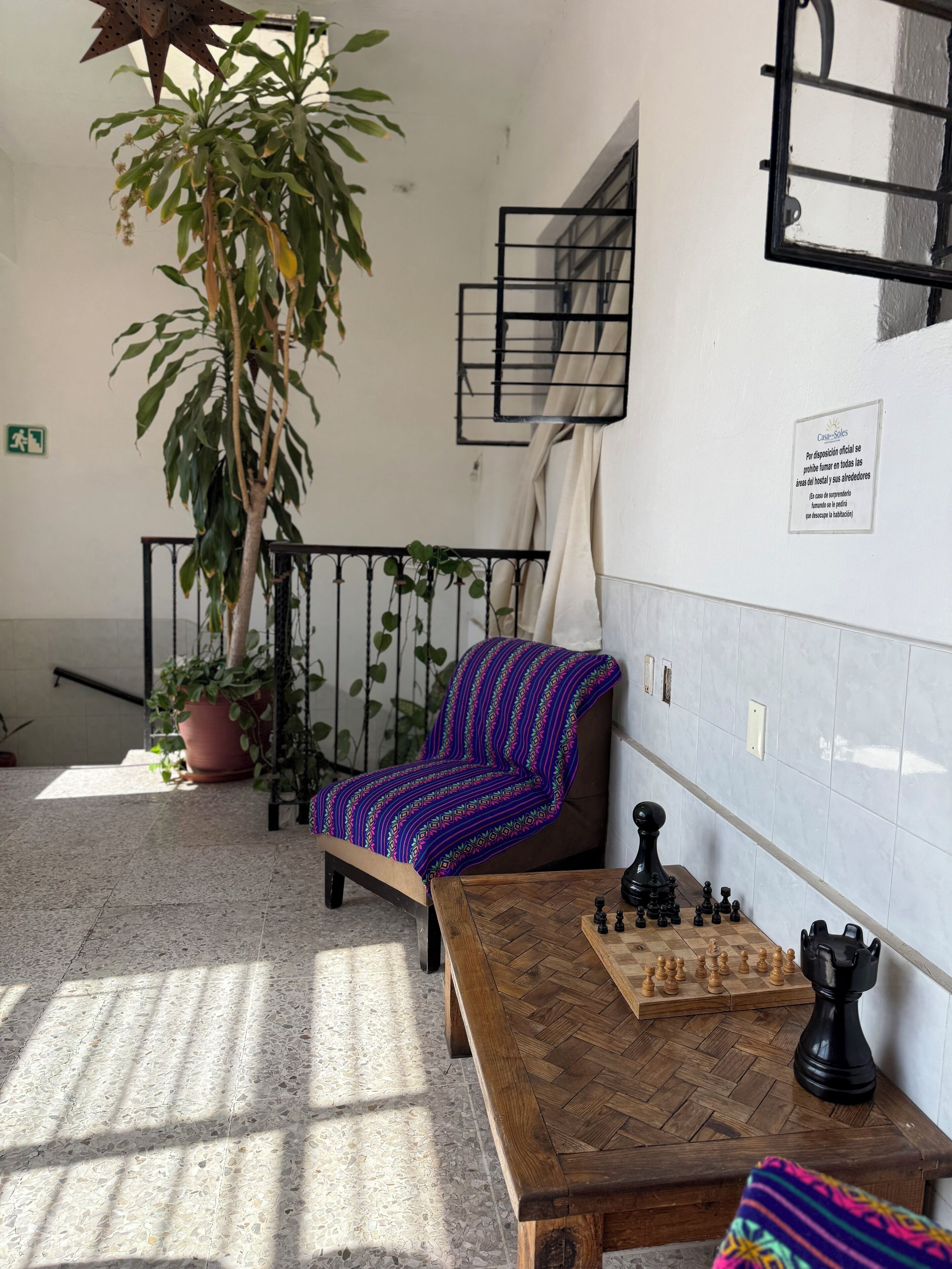 Una habitación con un tablero de ajedrez, una silla con una manta colorida, una planta en maceta y un cartel en la pared.