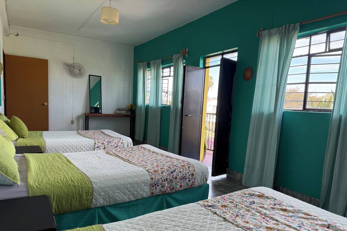 A room with two beds, a desk, a chair, a fan, and a window with curtains.