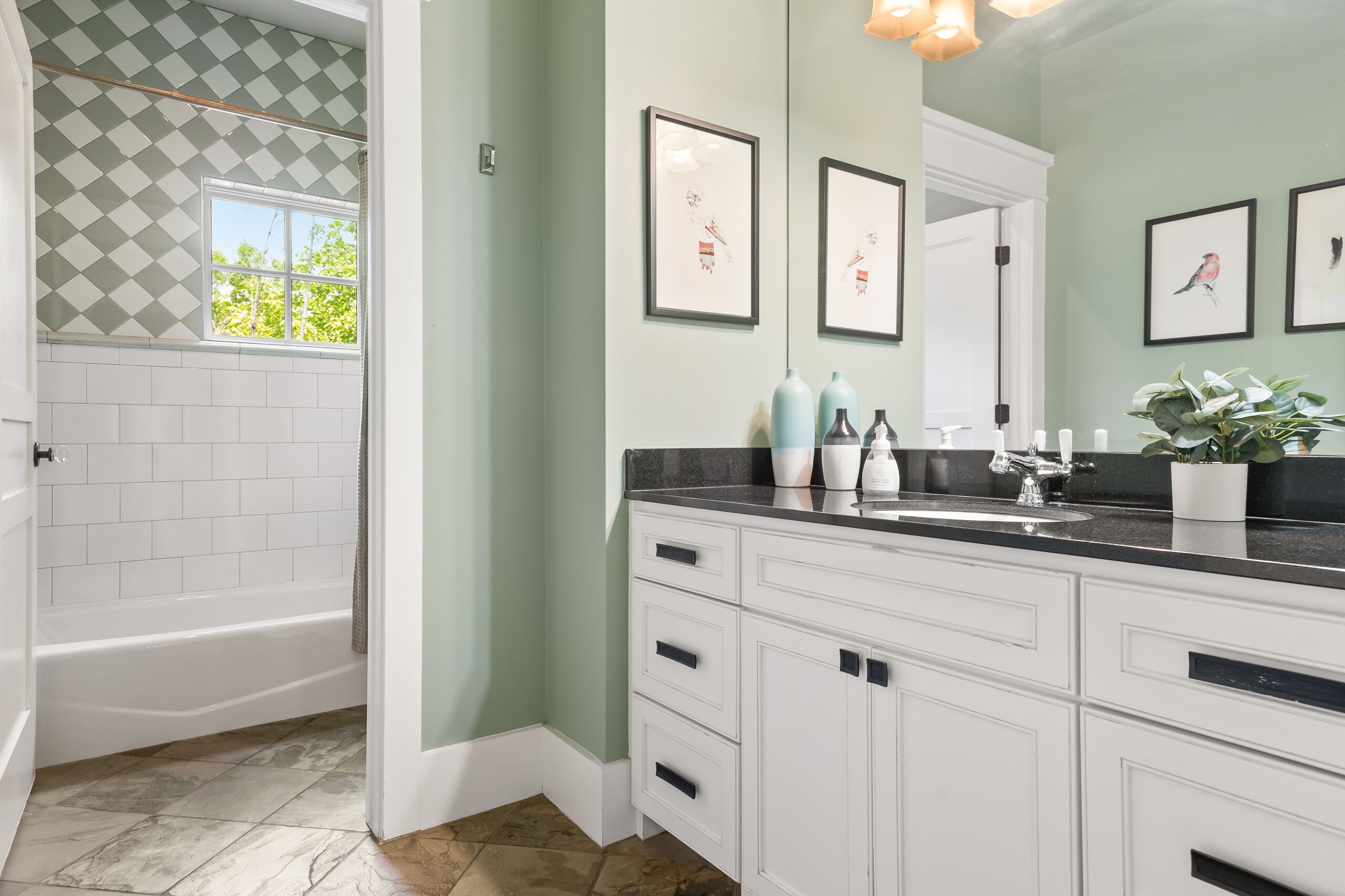 Un baño con bañera, armarios blancos, un lavabo con espejo, cuadros en la pared y una ventana con vistas a árboles.