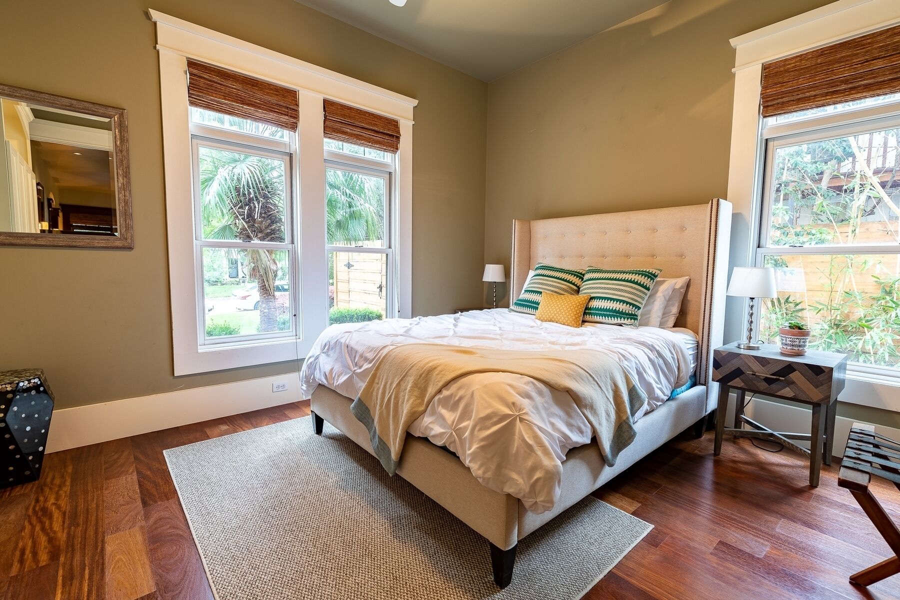 A bedroom with a bed, two windows with blinds, a bedside table with a lamp, and a view of greenery outside.