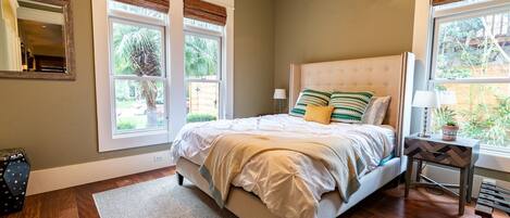 A bedroom with a bed, two windows with blinds, a bedside table with a lamp, and a view of greenery outside.