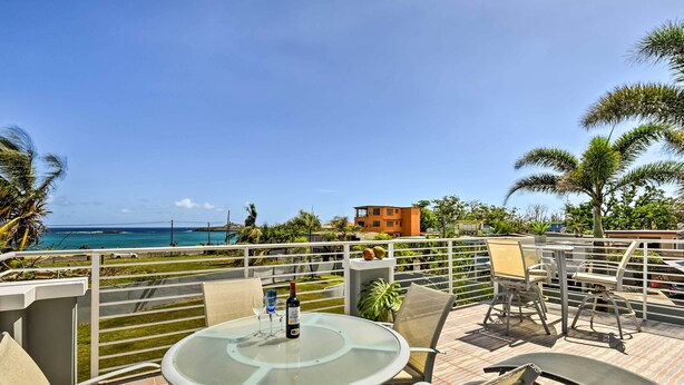 A terrace with a glass table, chairs, and a bottle of wine, overlooking a beach and ocean with palm trees and a building in the background.