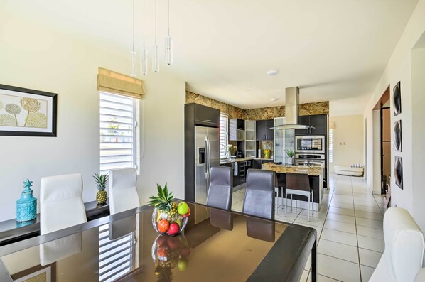 A modern kitchen with stainless steel appliances, a dining area with a glass table and white chairs, and a view of the city through the window.