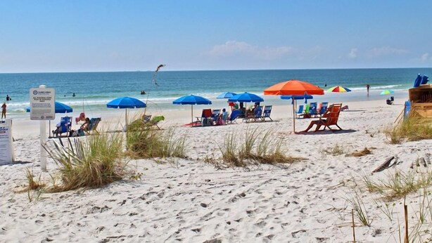 A beach with several blue and red umbrellas, lounge chairs, and a sign indicating a lifeguard on duty. - Cozy Beachfront Condo in Gulf Shores | Pool & Outdoor Grills | Sleeps 6 (Gulf Shores)