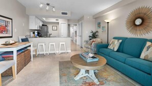 A modern living room with a teal sofa, a coffee table, and a kitchen in the background. - Cozy Beachfront Condo in Gulf Shores | Pool & Outdoor Grills | Sleeps 6 (Gulf Shores)