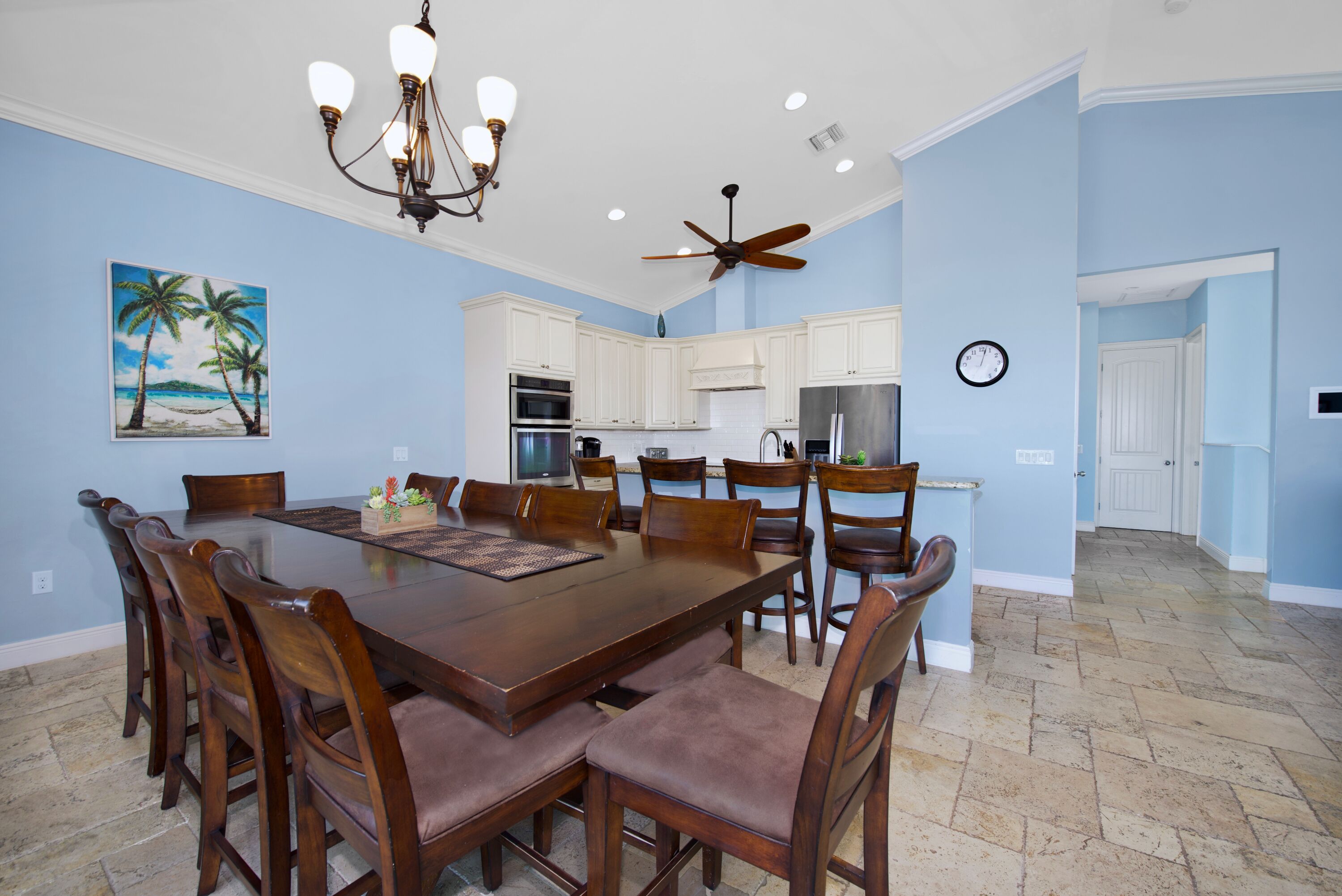 A spacious dining area with a large wooden table and matching chairs, a ceiling fan, and a kitchen area with white cabinets and appliances.