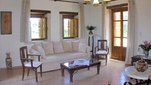 Ein geräumiges Wohnzimmer mit einer Couch, zwei Sessel, einem Couchtisch und einem Esstisch. Es gibt Fenster mit Vorhängen, eine Vase mit Blumen und ein Gemälde an der Wand.