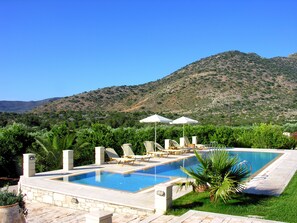 Une piscine entourée de chaises longues et de parasols, bordée de verdure et avec une chaîne de montagnes en toile de fond.