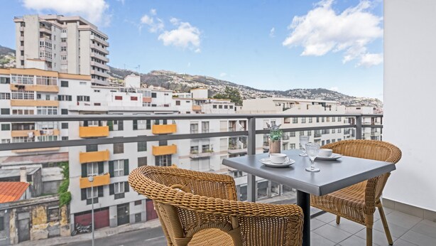 A balcony with wicker chairs and a table, overlooking a cityscape with buildings and a hill.