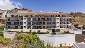 A multi-story residential building with balconies, a flat roof, and a hillside backdrop.