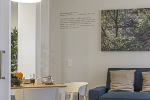 Una habitación con zona de comedor que incluye una mesa con un cuenco de fruta, una jarrón y vasos. Se ve un sofá azul y una silla blanca. En la pared hay un cartel informativo enmarcado sobre un sendero local.