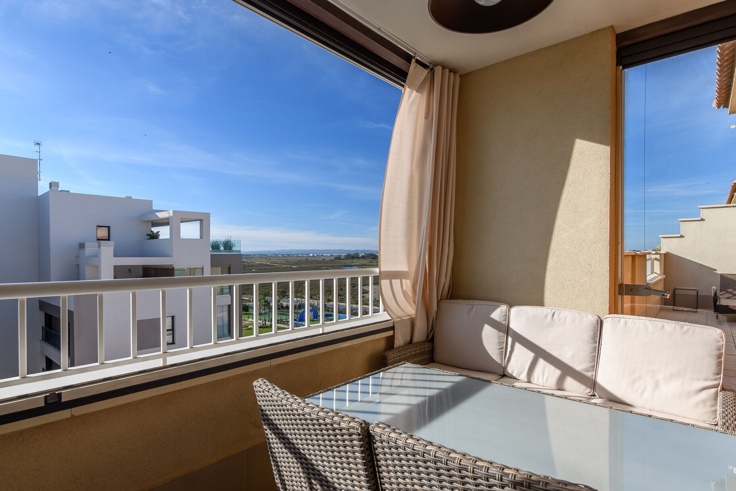 Un balcon doté d’une table en verre et de chaises en osier, offrant une vue sur les bâtiments et un ciel dégagé.