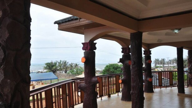 A covered outdoor area with wooden pillars and a metal railing, overlooking a beach and palm trees. - Villa D' Coco (Accra)