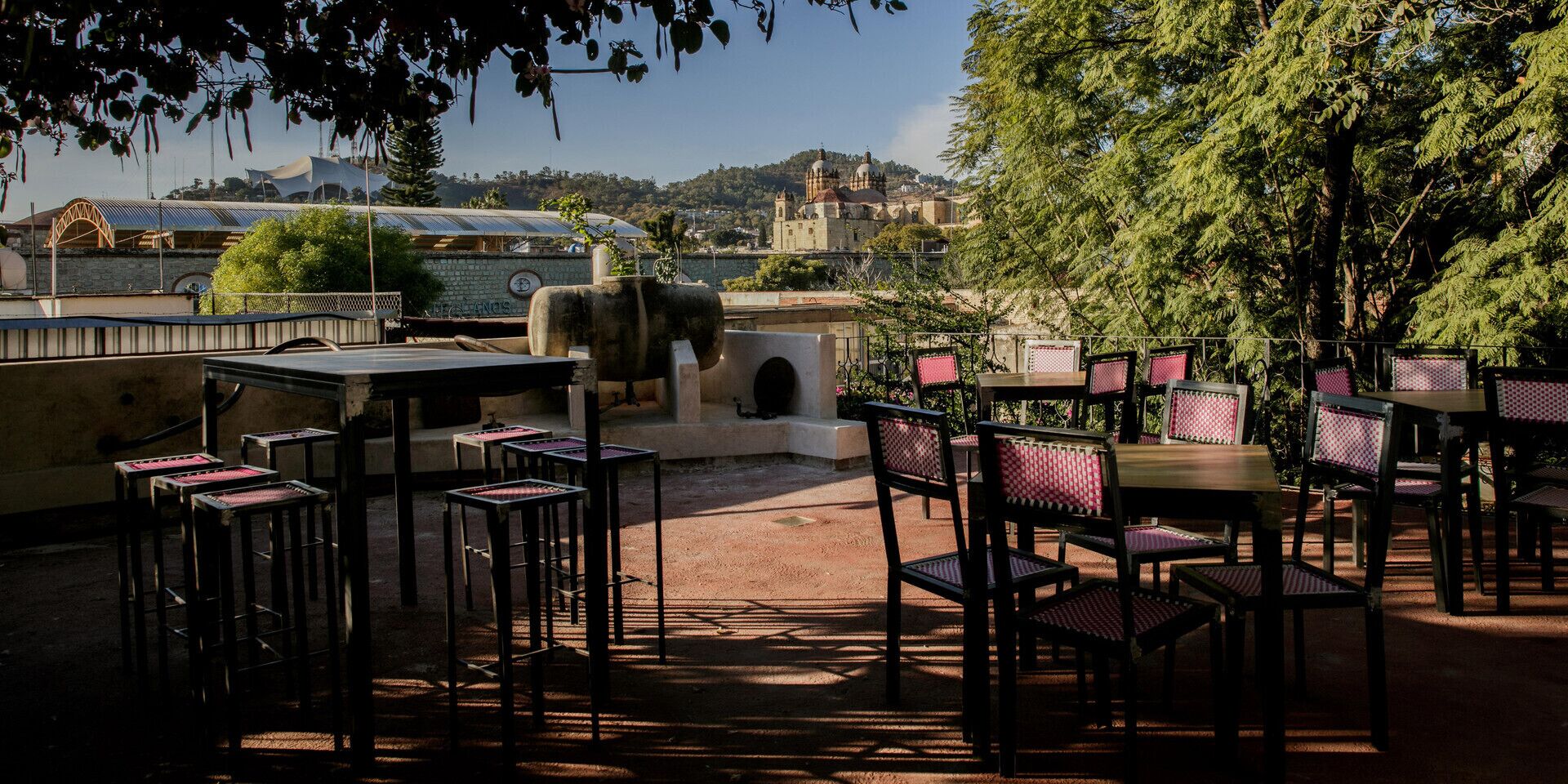 Un patio al aire libre con mesas y sillas, un muro de piedra y vistas a un paisaje urbano con edificios y una colina al fondo.